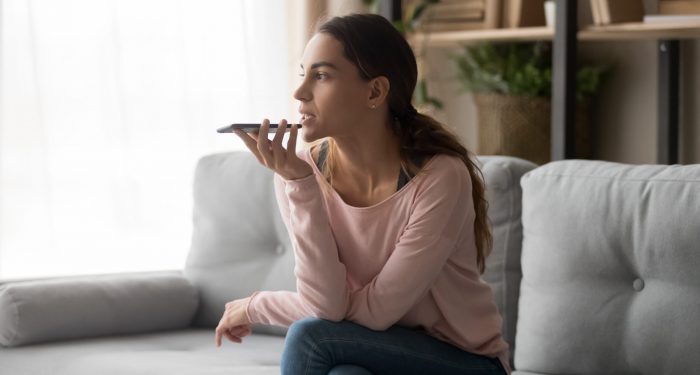 Serious young woman holding phone using mobile voice recognition or digital assistance app speak activate virtual assistant, record message or talk on speakerphone on smartphone sit on sofa at home