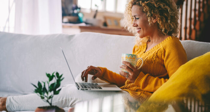 Real life of woman using laptop computer smilig and drinking a coffee at home comfortably sitting on the sofa. Modern female people use computer notebook in indoor leisure activity. Technology