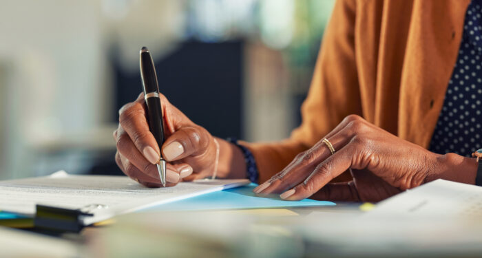 Close up of african american business woman hands writing on report while working in office. Successful black woman signing documents at home using a pen. Mid adult freelancer signing contract document on table.